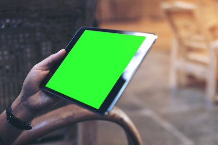 Mockup image of woman's hands holding black tablet pc with blank green screen on thigh in modern cafeの写真素材