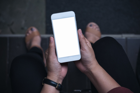 Top view mockup image of a woman sitting and holding white mobile phone with blank screen with concrete floor backgroundの写真素材