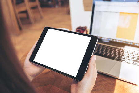 Mockup image of hands holding black tablet pc with white blank screen and laptop on wooden table background in cafeの写真素材