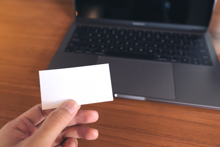 A hand holding white empty business card with laptop and coffee cup on wooden table in officeの写真素材