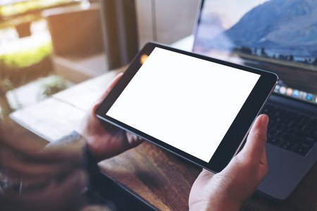 Mockup image of hands holding black tablet pc with white blank screen and laptop on wooden table background in cafeの写真素材