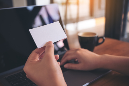 A hand holding white empty business card while using laptop with coffee cup on wooden table in officeの写真素材