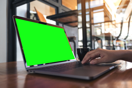 Mockup image of a hand using and typing on laptop with blank green screen and coffee cup on wooden table in cafeの写真素材