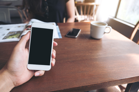Mockup image of a man's hand holding white mobile phone with blank black screen , white coffee cups and woman reading book in modern loft cafeの写真素材