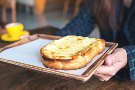 Closeup image of a woman holding a piece of sliced bread toast with cheese in wooden plateの写真素材
