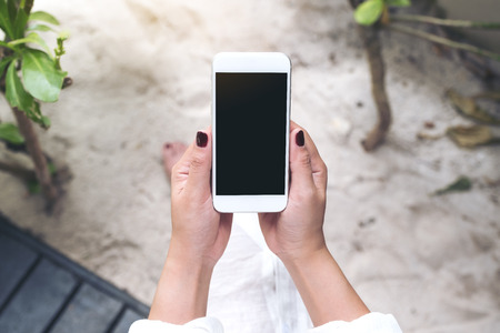 Top view mockup image of a woman's hand holding white mobile phone with blank black desktop screen with sand and beach backgroundの写真素材