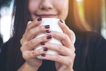Closeup image of a beautiful Asian woman holding a white cup of hot coffee with feeling happy in cafeの写真素材