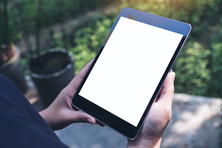 Mockup image of woman's hands holding black tablet pc with blank white screen and green nature backgroundの写真素材