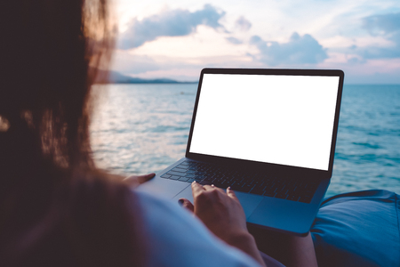 Mockup image of a woman using laptop with blank white desktop screen while sitting by the sea with blue sky backgroundの写真素材