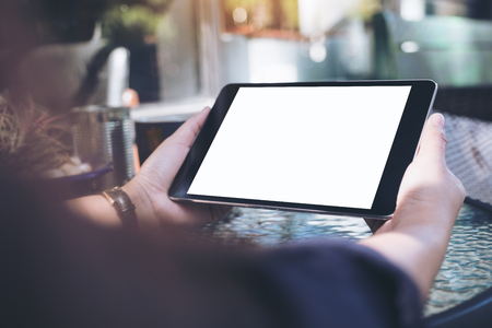 Mockup image of hands holding black tablet pc with blank white desktop screen and coffee cup on glass tableの写真素材