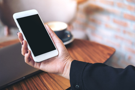 Mockup image of a woman's hand holding white mobile phone with blank black desktop screen and laptop on wooden table in cafeの写真素材