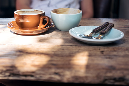 Two cups of hot coffee and a plate with spoon and fork on vintage wooden table with people in...