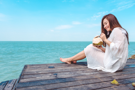 A happy beautiful asian woman on white dress sitting at the wooden terrace and drinking coconut juice with sea and blue sky backgroundの写真素材