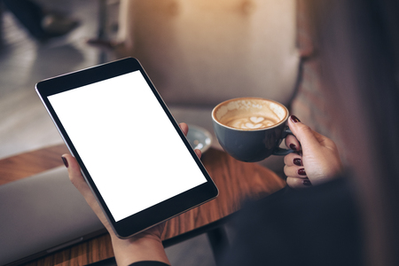 Mockup image of woman's hands holding black tablet pc with blank white desktop screen and coffee cup in cafe backgroundの写真素材