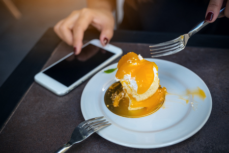 Closeup image of a woman using smartphone while cutting and eating an orange cake with fork on white plate on wooden table in cafeの写真素材