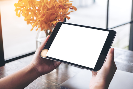 Mockup image of hands holding black tablet pc with blank white screen and flower vase on wooden table in cafeの写真素材