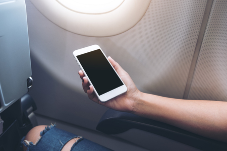 Mockup image of woman's hands holding a white smart phone with blank black desktop screen next to an airplane windowの写真素材