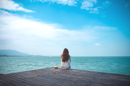 A beautiful asian woman on white dress sitting and looking at the sea and blue sky on wooden balcony with feeling relaxedの写真素材
