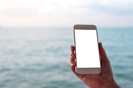Mockup image of a hand holding and showing white mobile phone with blank black desktop screen in front of the sea and blue sky backgroundの写真素材