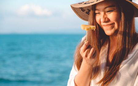 Portrait image of a happy beautiful asian woman enjoy eating pineapple by the sea with clear blue sky backgroundの写真素材