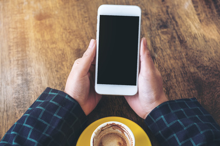 Top view mockup image of hands holding white mobile phone with blank black desktop screen and yellow coffee cup on wooden table in modern cafeの写真素材