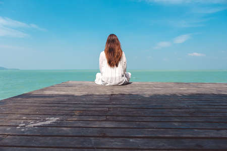 A beautiful asian woman on white dress sitting and looking at the sea and blue sky on wooden balcony with feeling relaxedの写真素材