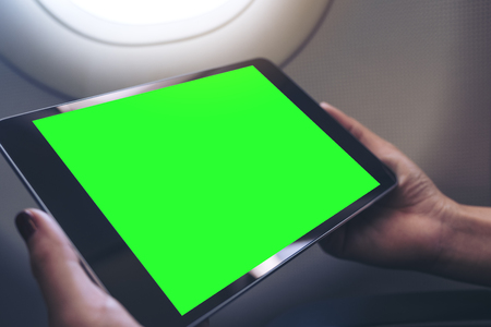 Mockup image of a woman holding and looking at black tablet pc with blank green desktop screen next to an airplane windowの写真素材