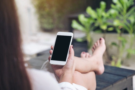 Mockup image of a woman sitting and holding white mobile phone with blank black desktop screen , feeling relaxed with sand and beach backgroundの写真素材