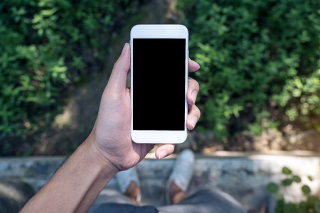 Mockup image of a man's hand holding white mobile phone with blank black screen while standing on concrete polishing floor with green leave and nature backgroundの写真素材