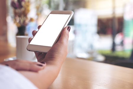 Mockup image of a woman's hand holding white mobile phone with blank desktop screen and coffee cup in cafeの写真素材