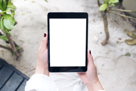 Mockup image of a woman's hand holding black tablet pc with blank white desktop screen with sand and beach backgroundの写真素材