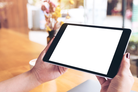 Mockup image of woman's hands holding black tablet pc with blank white desktop screen , laptop and coffee cup on wooden table in cafeの写真素材