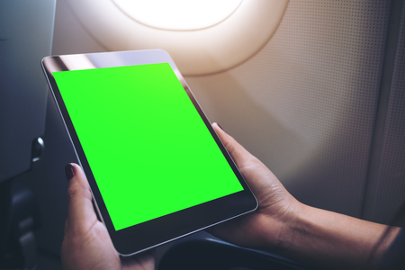 Mockup image of a woman holding and looking at black tablet pc with blank green desktop screen next to an airplane windowの写真素材