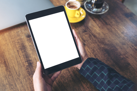 Mockup image of hands holding black tablet pc with blank desktop white screen and coffee cup on wooden table in cafe backgroundの写真素材