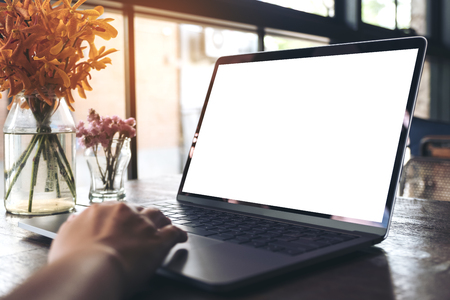 Mockup image of a hand using and touching laptop with blank white desktop screen and flower vase on wooden table in cafeの写真素材