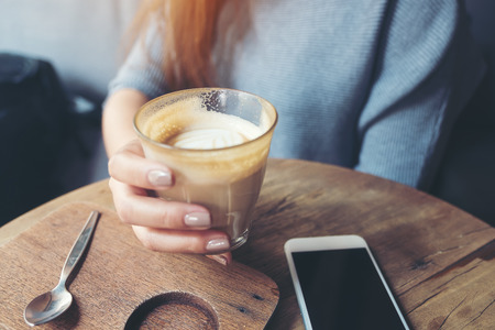 Closeup image of a woman's hand holding a cup of coffee with smart phone  on wooden table in cafeの写真素材