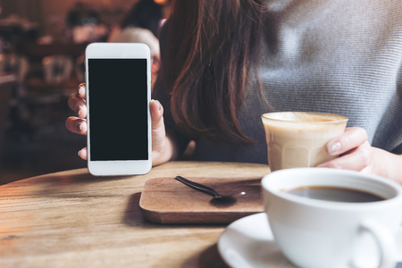 Mockup image of a woman holding and showing white mobile phone with blank black desktop screen with coffee cups on vintage wooden tableの写真素材