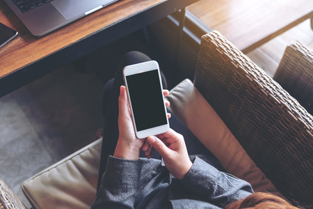 Mockup image of a woman's hands holding white mobile phone with blank black screen on thighの写真素材