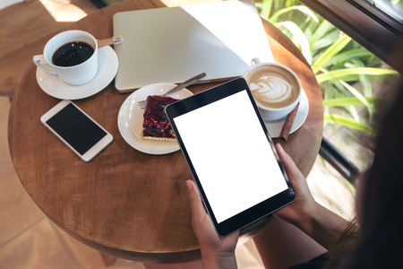 Top view mockup image of hands holding black tablet pc with blank white screen , smart phone , laptop , coffee cup and cake on wooden table in cafeの写真素材
