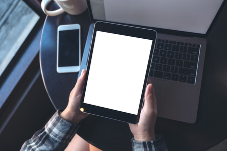 Top view mockup image of hands holding black tablet pc with white blank screen and laptop , coffee cup on wooden table background in cafeの写真素材