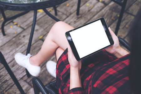 Mockup image of woman's hands holding black tablet pc with blank white screen on thigh with wooden floor background in vintage cafeの写真素材