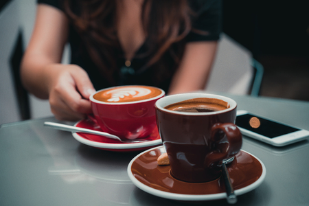 Closeup image of a woman's hand holding latte coffee cup with Americano coffee cup and smartphone on glass table in cafeの写真素材