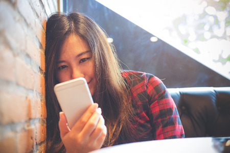 Closeup image of a smiley beautiful Asian woman leaning on brick wall while using and looking at smart phone in modern cafeの写真素材
