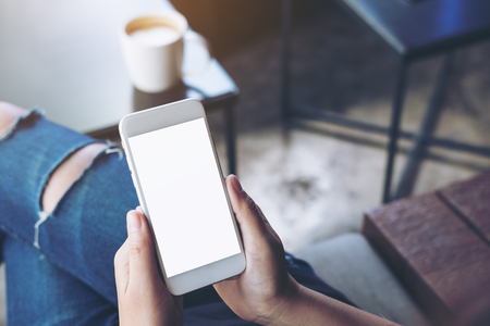 Mockup image of woman's hands holding white mobile phone with blank screen on thigh and coffee cup on the tableの写真素材