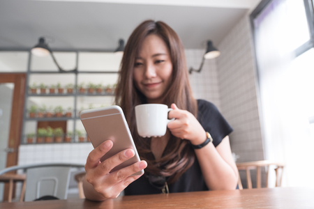 Closeup image of a beautiful Asian woman with smiley face holding and using smart phone while drinking hot coffee on wooden table in modern cafeの写真素材