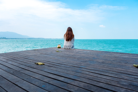 A beautiful asian woman on white dress sitting and looking at the sea and blue sky on wooden balcony with feeling relaxedの写真素材