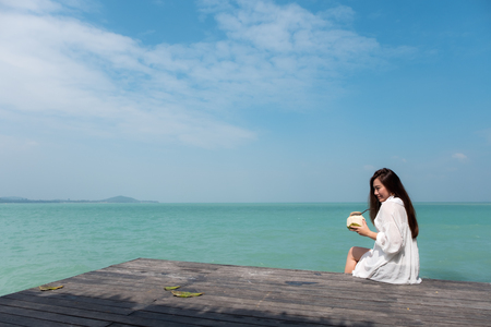 A beautiful asian woman on white dress sitting at the terrace drinking coconut juice with sea and blue sky background with feeling relaxedの写真素材