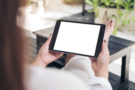 Mockup image of a woman sitting and holding black tablet pc with blank white desktop screen , feeling relaxed with sand and beach backgroundの写真素材