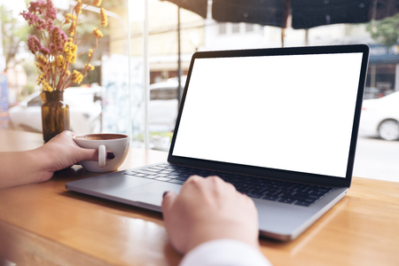 Mockup image of a businesswoman's hand using laptop with blank white desktop screen while drinking hot coffee on wooden table in cafeの写真素材