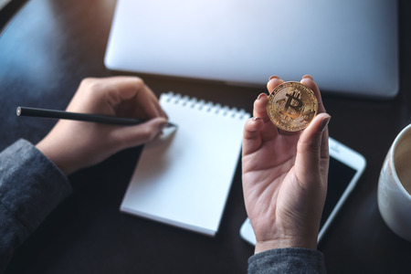 Topview image of a woman's hands holding bitcoin and writing on empty notebook with laptop and mobile phone on the tableの写真素材